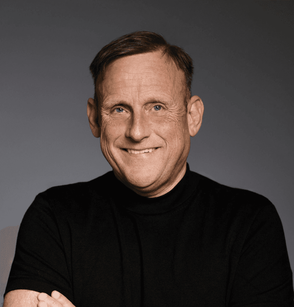 Mann mit schwarzem Pullover und verschränkten Armen in die Kamera lächelnd.