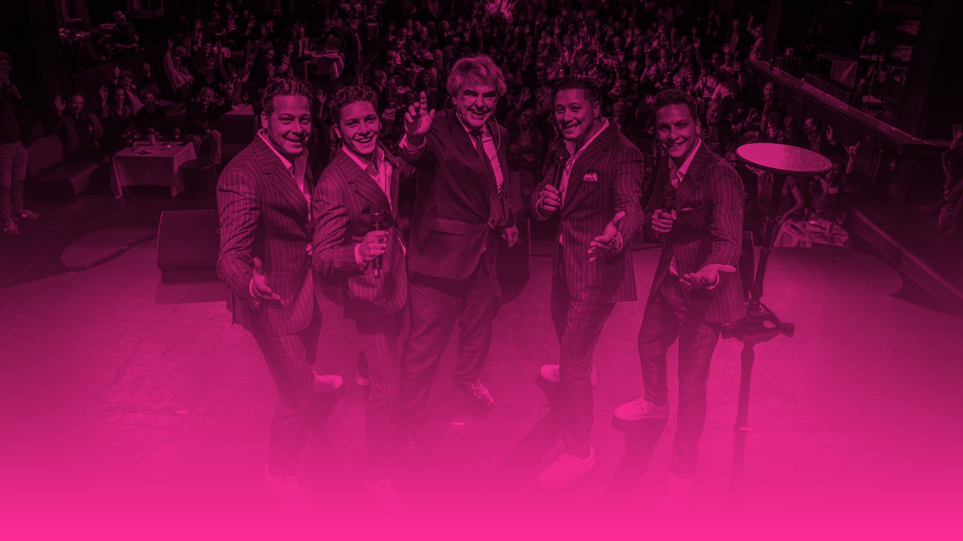 Das Foto zeigt die gleiche Szene wie das allererste Bild, das du mir geschickt hast: Eine fünfköpfige Männergruppe steht auf einer Bühne vor einem vollen, jubelnden Publikum. Ein älterer Mann im dunklen Anzug steht im Zentrum und zeigt mit dem Finger nach oben, flankiert von vier jüngeren Männern in identischen Nadelstreifenanzügen und weißen Turnschuhen, die Mikrofone halten. Der entscheidende Unterschied bei dieser Version: Das gesamte Bild ist stark verfremdet und mit einem intensiven magentafarbenen (pinken) Filter belegt. Wie bei den vorherigen Bildern dieser gestalterischen Serie geht das untere Drittel des Fotos fließend in eine durchgehende, leuchtend pinke Farbfläche über.
