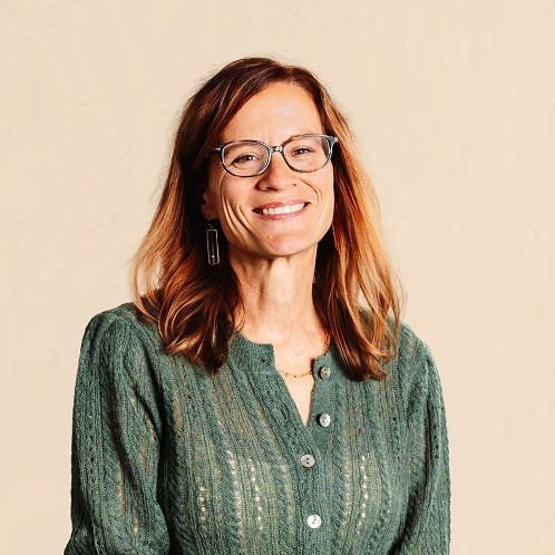 Smiling woman wearing glasses and a green textured blouse, standing against a neutral background.