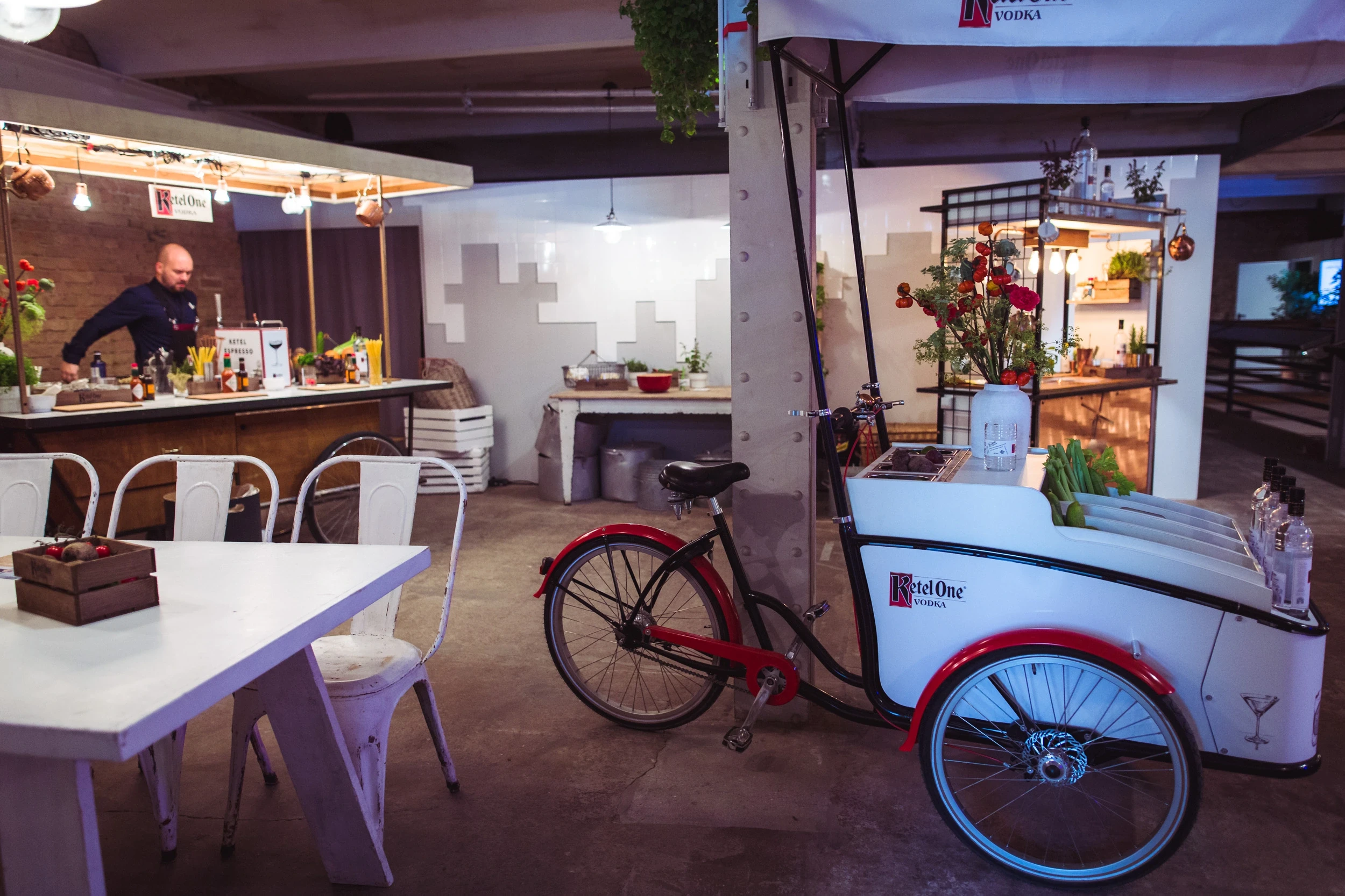 Ketel One cargo bike bar with red wheels and white box at DIAGEO BCB18, next to a white bar with bartender and market-stall canopies.