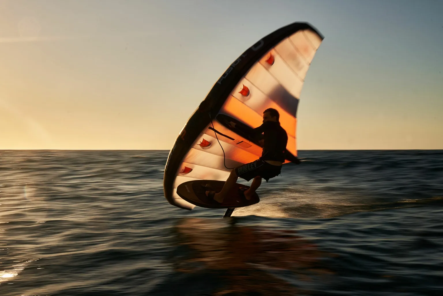 Sunset wingfoiling session with rider gliding above ocean surface, showcasing dynamic water sport action and Ozone Wing performance