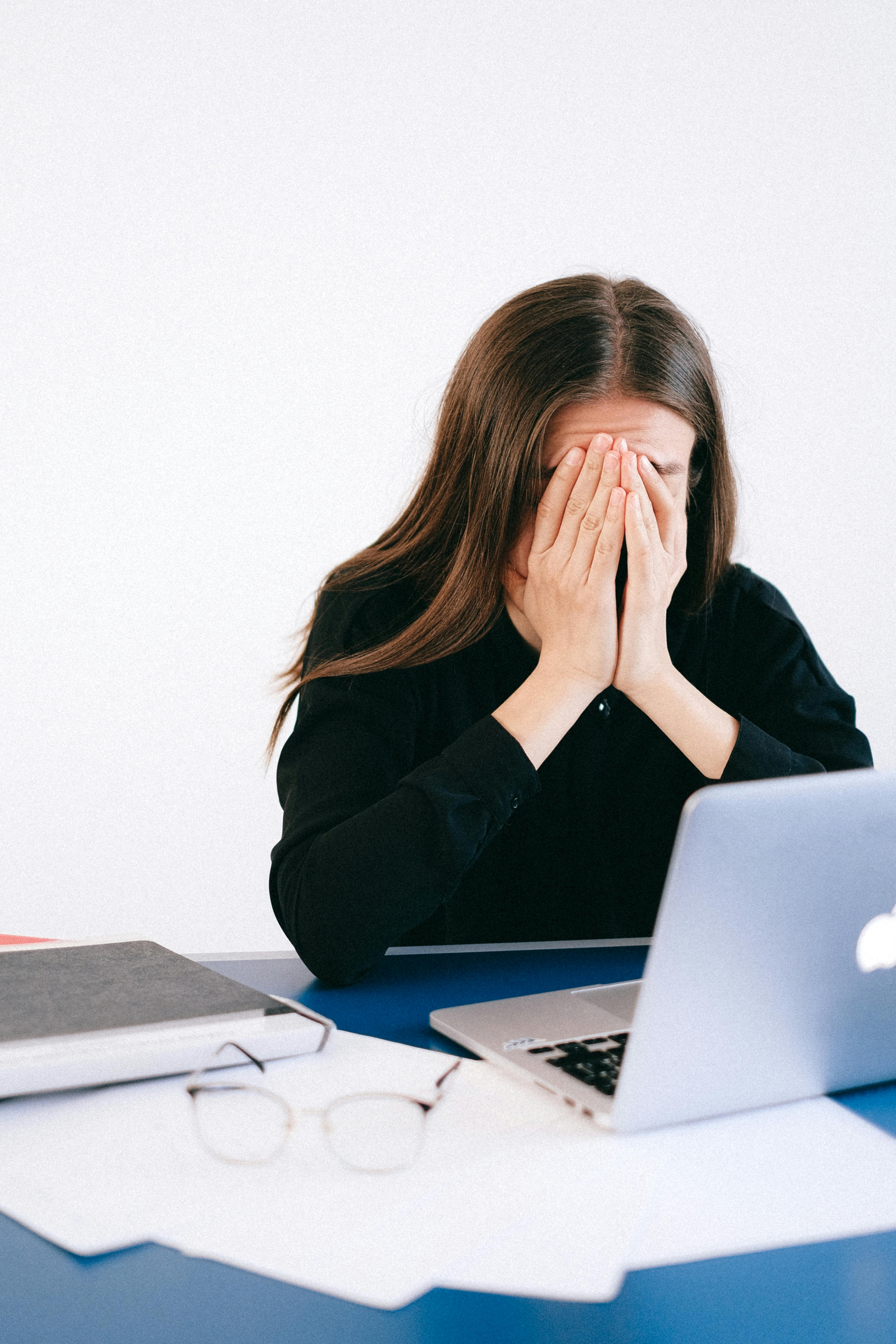 Woman sitting at a desk with a laptop, covering her face with her hands in frustration.