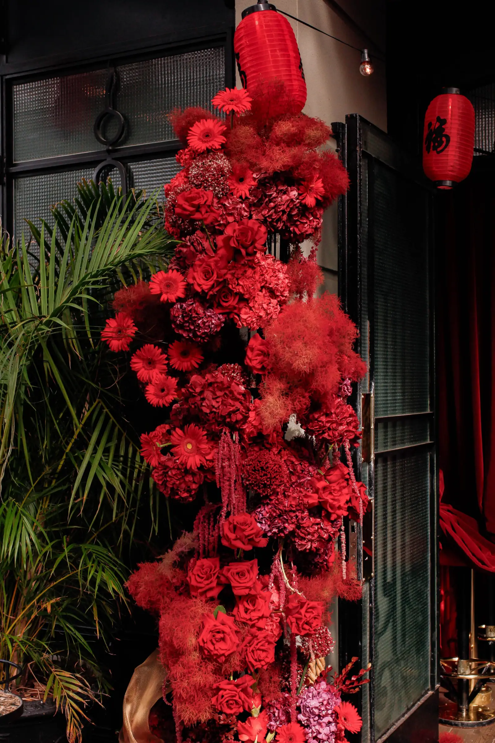 Columna floral alta de rosas, hortensias y plumeros rojos junto a faroles orientales.