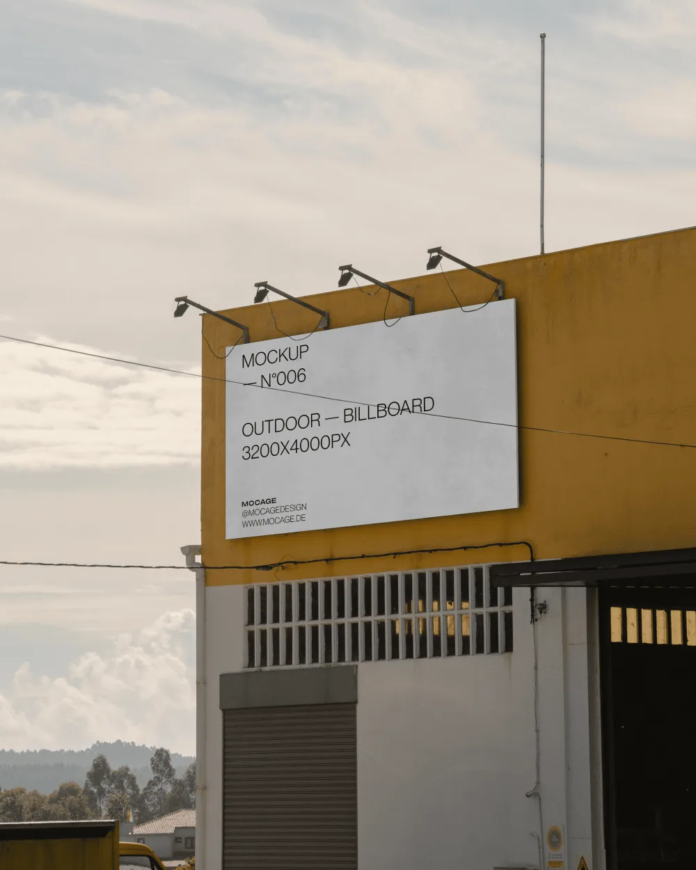 Outdoor billboard mockup displayed on a yellow building with industrial features, illuminated by overhead spotlights. The cloudy sky in the background adds depth to the scene.