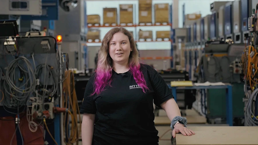Auszubildende Lena von RITTER Starkstromtechnik steht in der Ausbildungswerkstatt und blickt in die Kamera. Im Hintergrund sind Schaltschränke und Arbeitsplätze der Fertigung zu sehen.