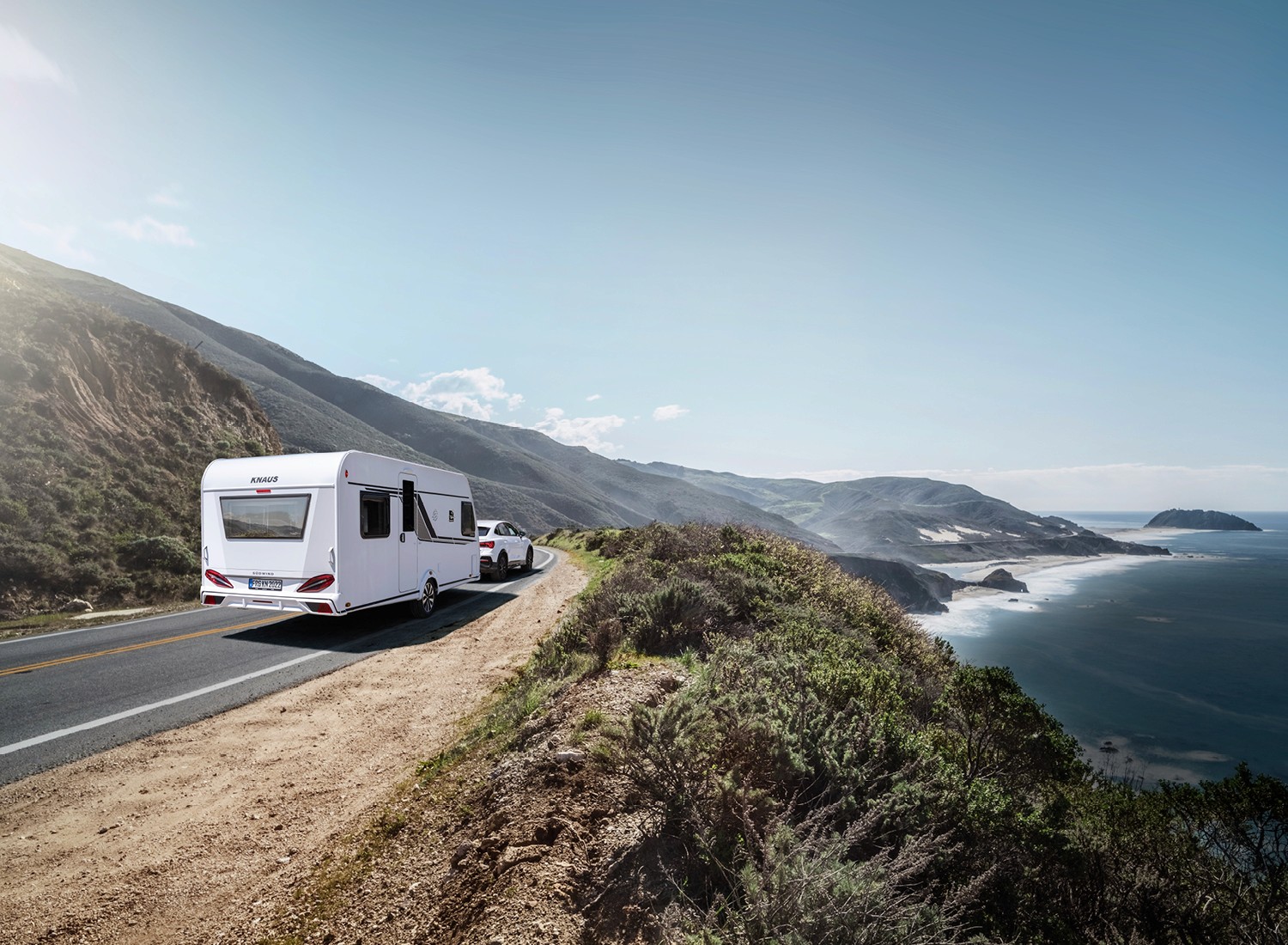 Ein KNAUS Wohnwagen wird von einem Auto entlang einer malerischen Küstenstraße gezogen – mit Blick auf das Meer, die Felsküste und die sonnenbeschienenen Hügel im Hintergrund.
