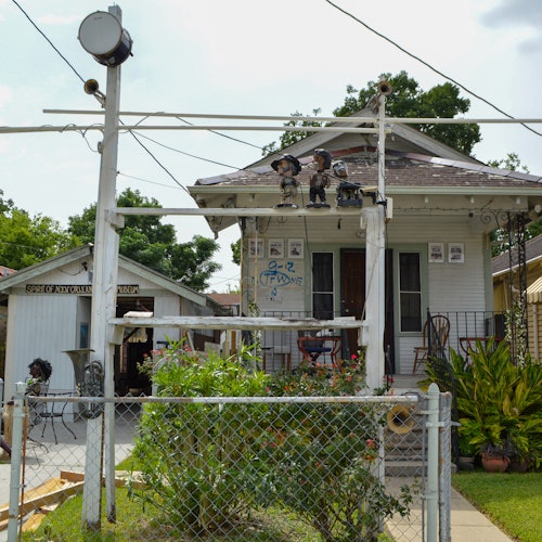 在前院有装饰物的小房子，有一个金属栅栏，还有一个写着“5-C's JAZZ”的牌子，周围环绕着郁郁葱葱的绿色植物。