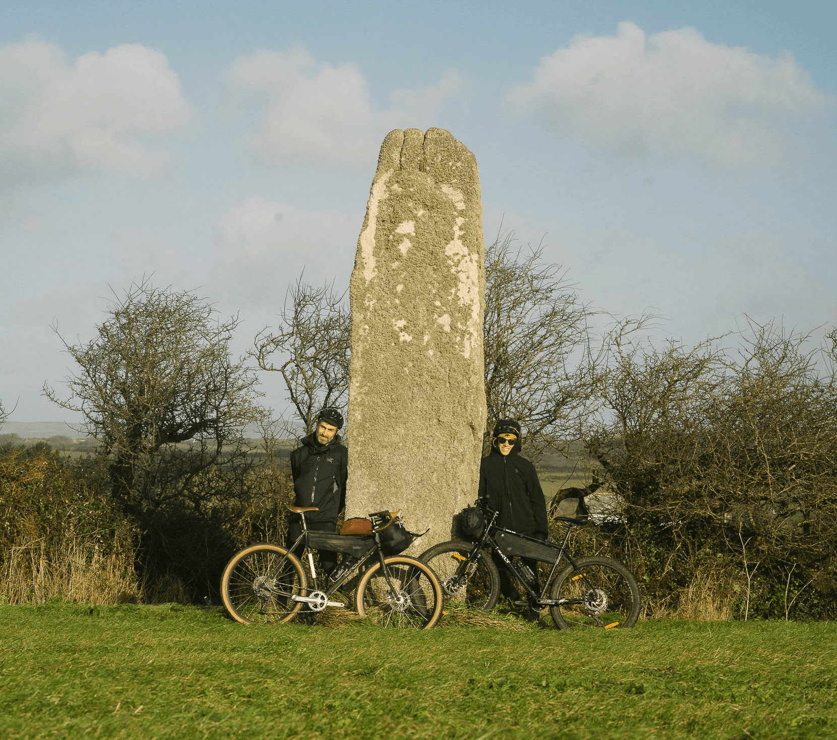 Two cyclists standing beside a stone monument in a rural landscape