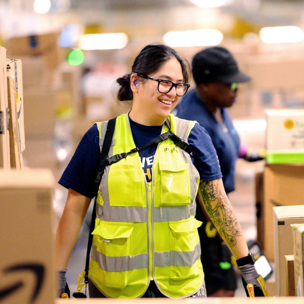A smiling woman wearing yellow safety vest in an Amazon warehouse filled with boxes.