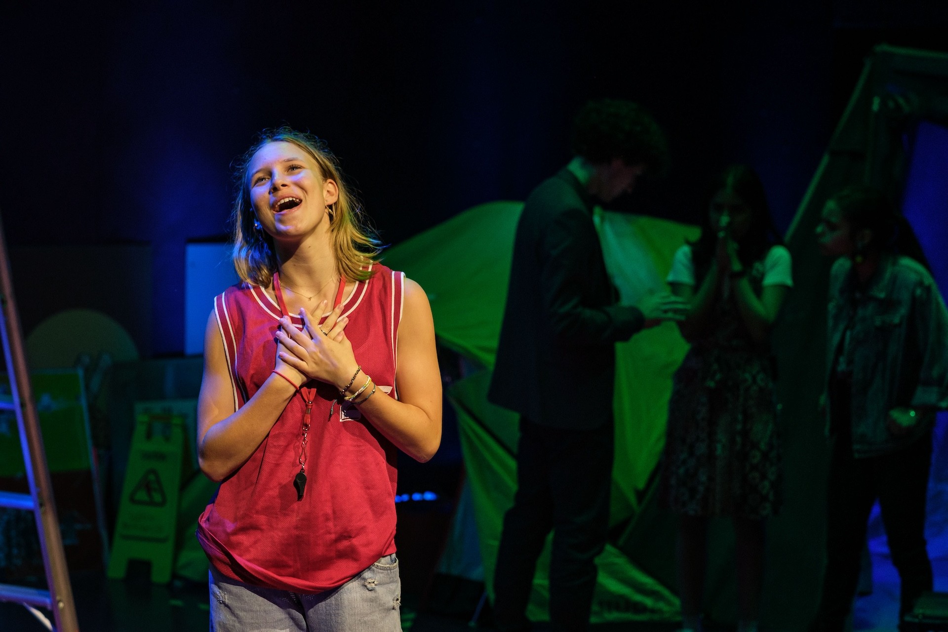A performer in a red sports singlet stands downstage with hands clasped to their chest, singing or speaking emotionally. In the background, other performers gather near a green tent under moody blue and green lighting.
