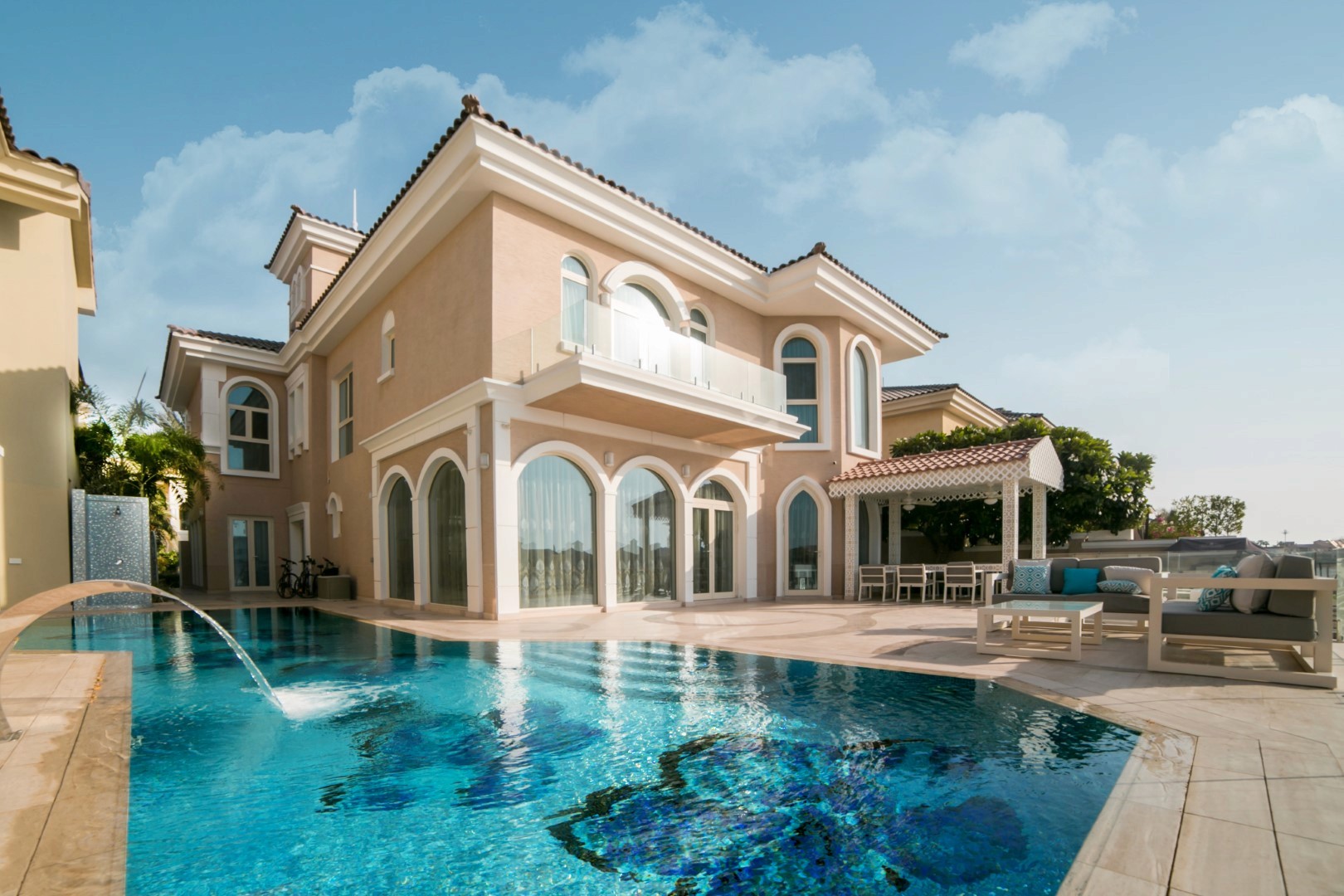 Luxurious Mediterranean-style villa with arched windows and a spacious terrace overlooking a serene swimming pool on a sunny day.