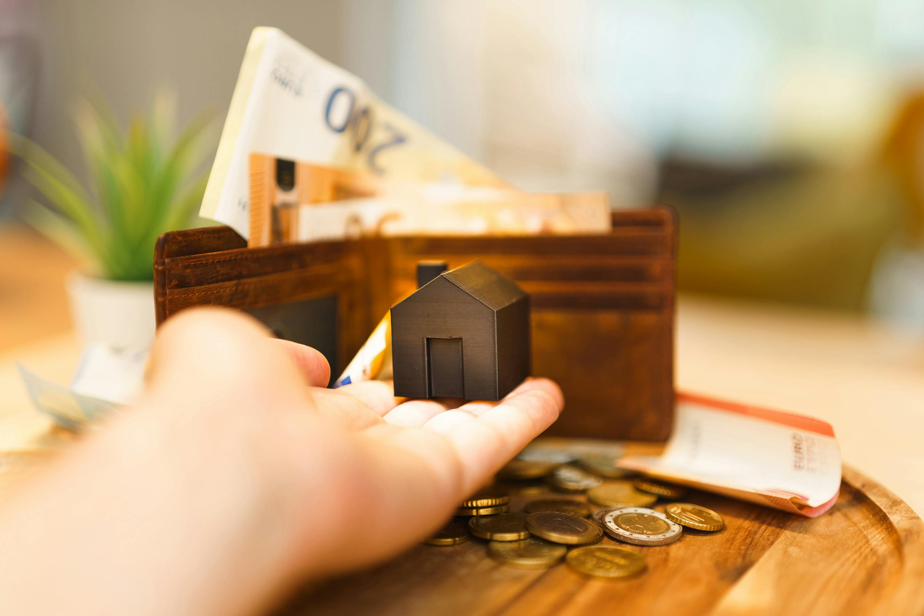 A hand reaches into a wallet containing cash and credit cards, set on a wooden table with a small plant.