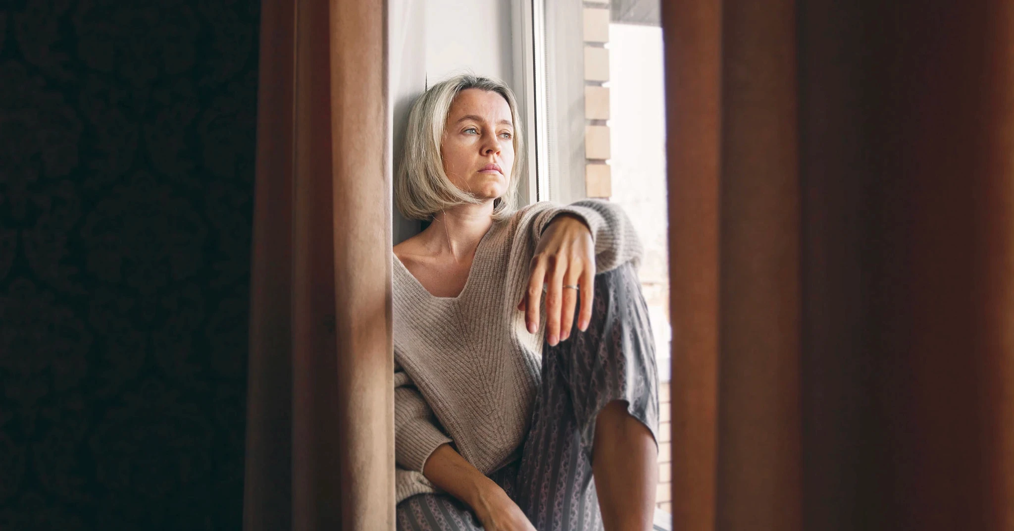 En kvinna sitter vid ett fönster och ser tankfull och ledsen ut.