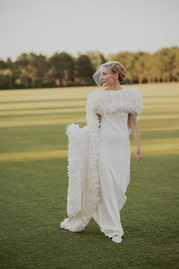 Novia con vestido blanco y capa texturada sobre campo abierto para sesiones editoriales que transmiten pureza, movimiento y sofisticación moderna.