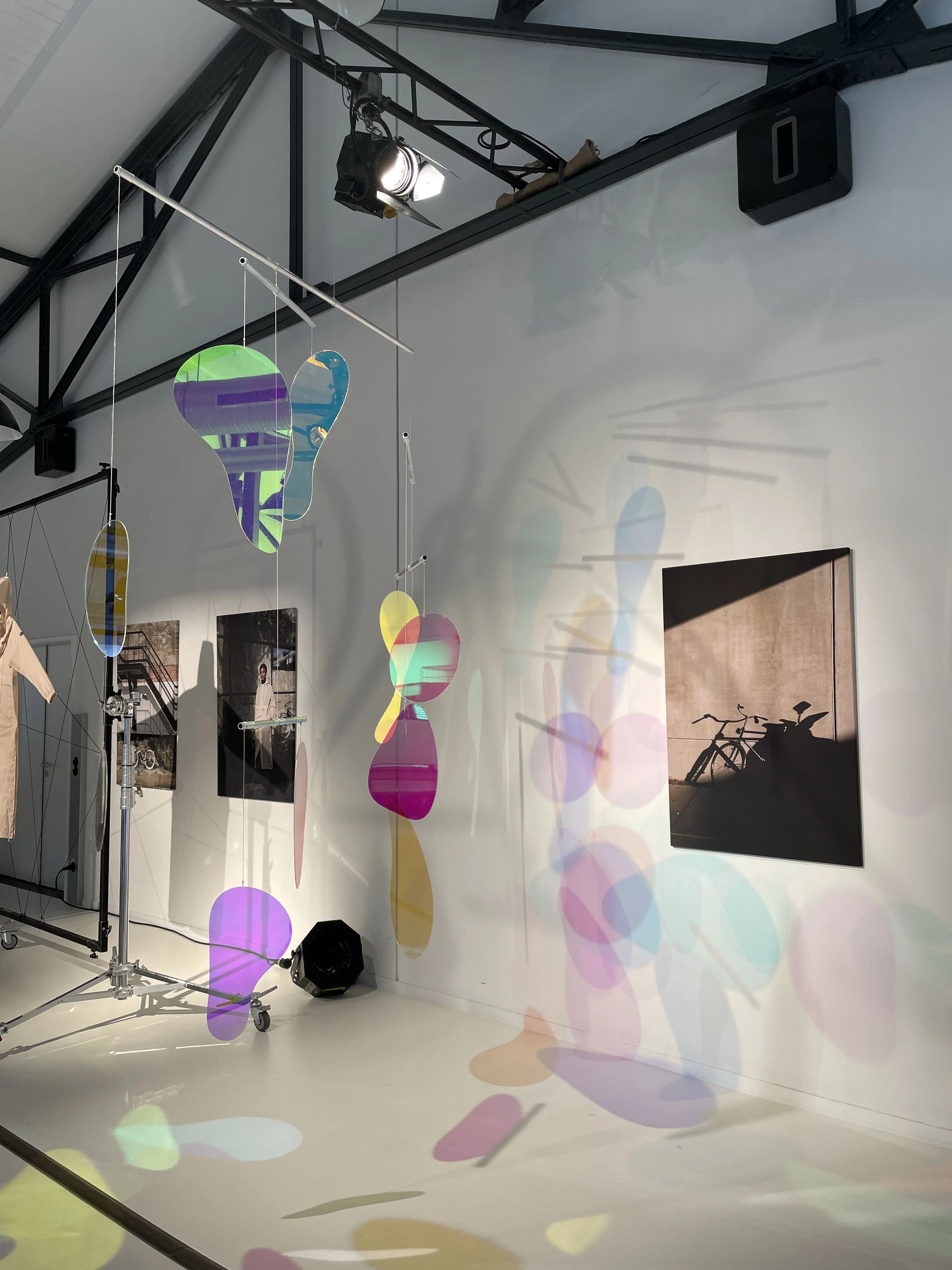 Iridescent acrylic mobiles hovering before black-and-white photography, throwing colorful light spots across the AGU gallery walls.