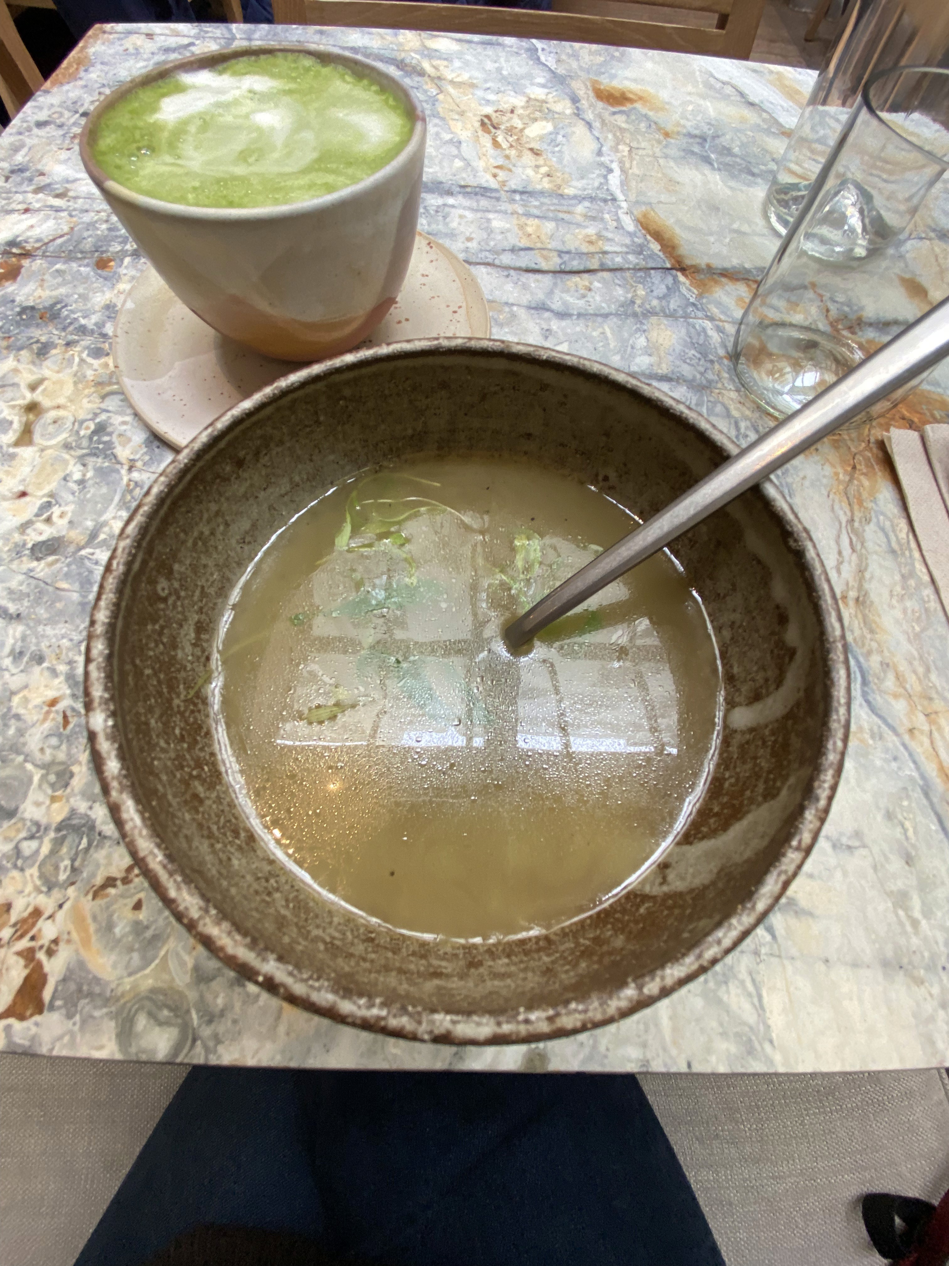 Un bol de soupe avec une cuillère et un matcha latte vert mousseux sur une table en marbre. Une carafe est visible.