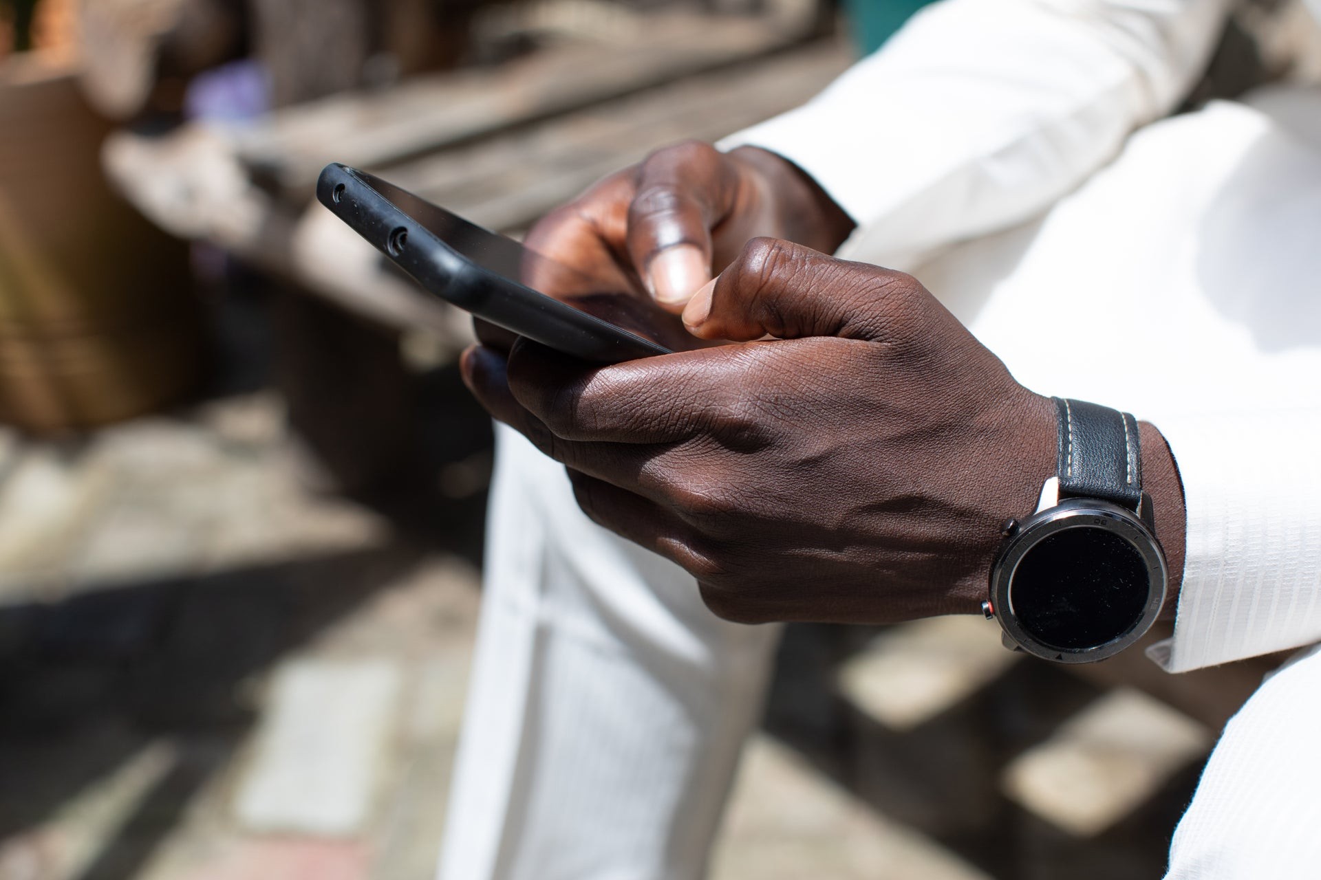 A close-up shot of a person's hands holding and typing on a smartphone with a black case. The individual has dark skin, is wearing a white long-sleeved garment, and has a black smartwatch with a leather strap on their wrist. The background is softly blurred, showing glimpses of an outdoor setting with wooden furniture and paved ground in natural sunlight.