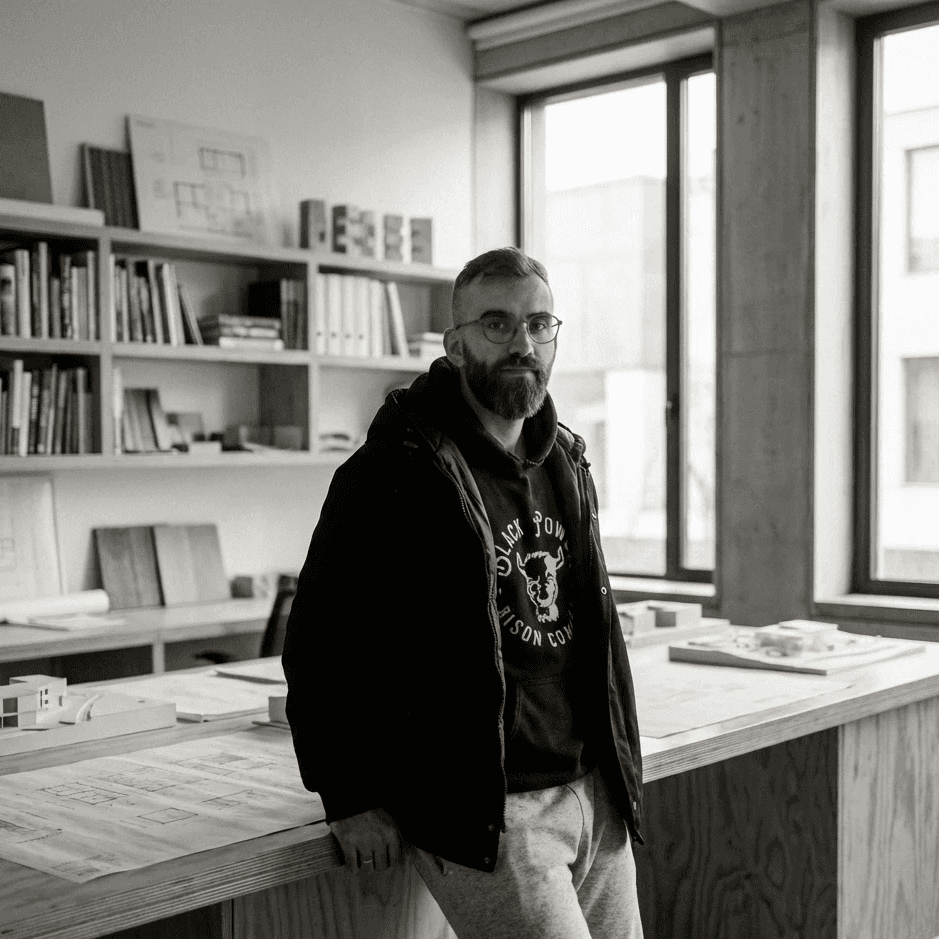 A man reading from a tablet in a collaborative office environment. Desks with documents and a discussion between colleagues are visible in the background.