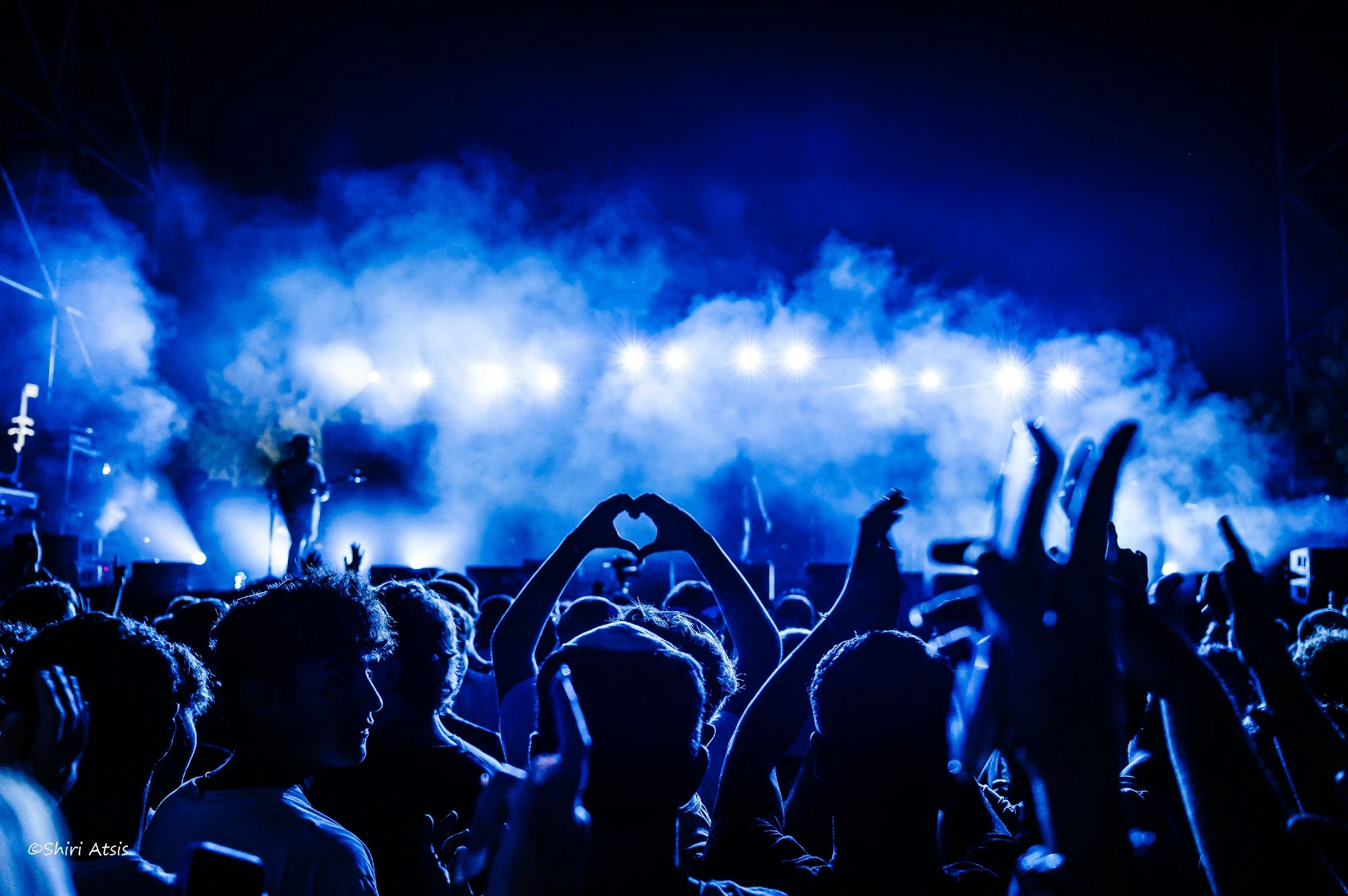 צילום אמנותי של קהל בהופעה חיה תחת תאורה כחולה עוצמתית, ידיים יוצרות צורת לב מול הבמה. צילום של הצלמת שירי עציץ Shiriz המתעד רגעי חיבור ורגש במוזיקה.