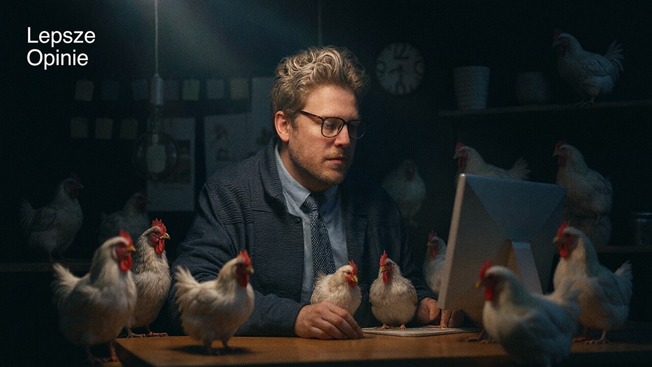 A man wearing glasses works at his computer in a modern office that is completely overrun with live chickens.