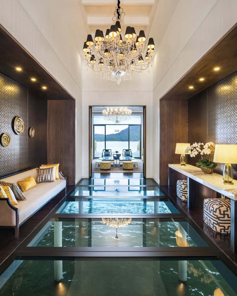 Interior view of The St. Regis, Langkawi, featuring elegant decor, a chandelier, a glass floor showcasing water underneath, and seating area overlooking the scenic landscape.