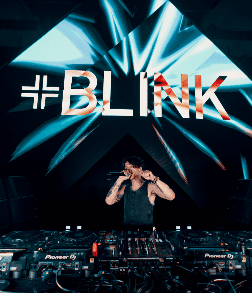 A DJ performing on stage with a microphone, surrounded by DJ equipment, as vibrant visuals are displayed in the background.