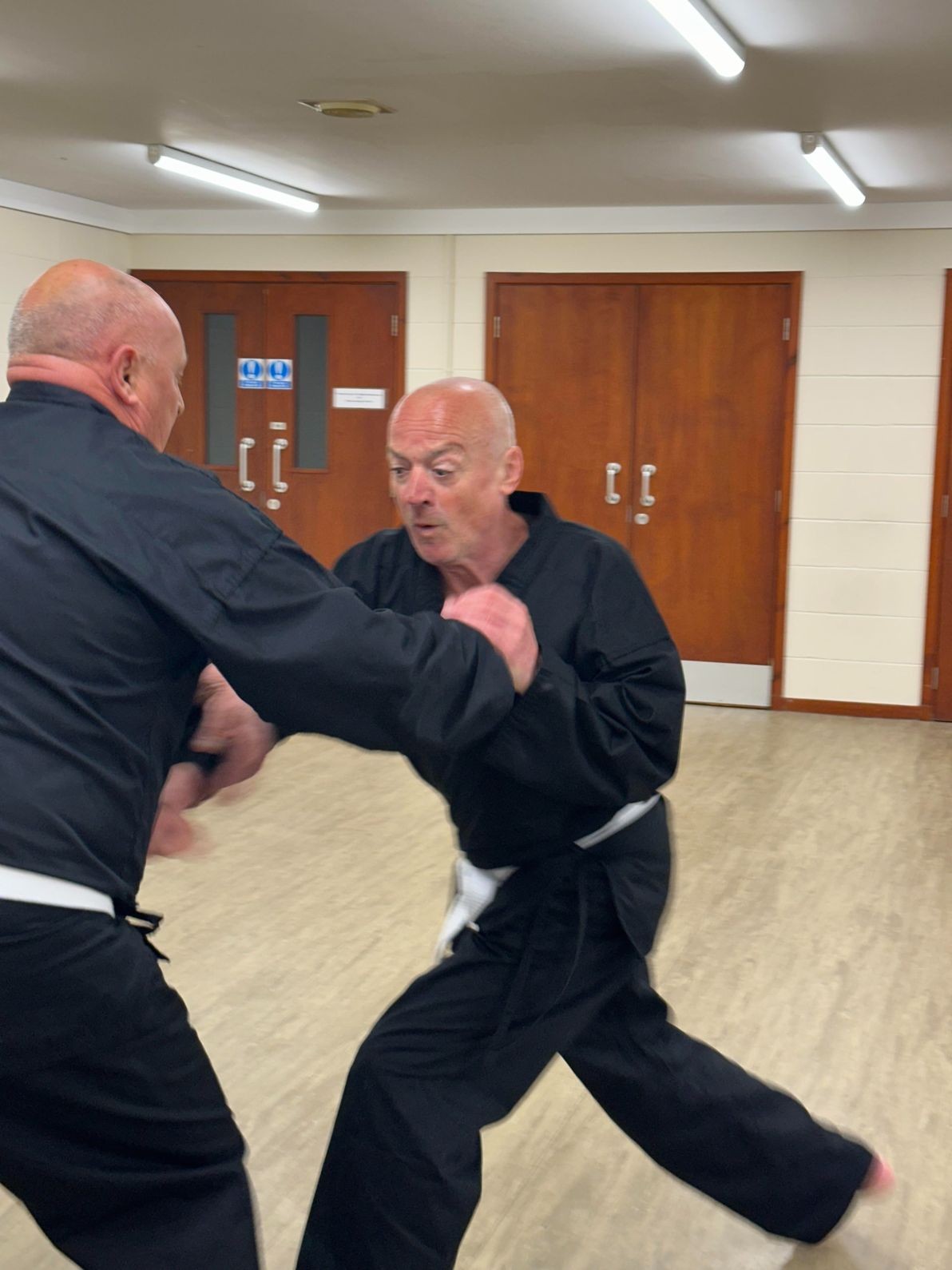 Two men in black martial arts uniforms practice sparring in a brightly lit room with wooden doors.