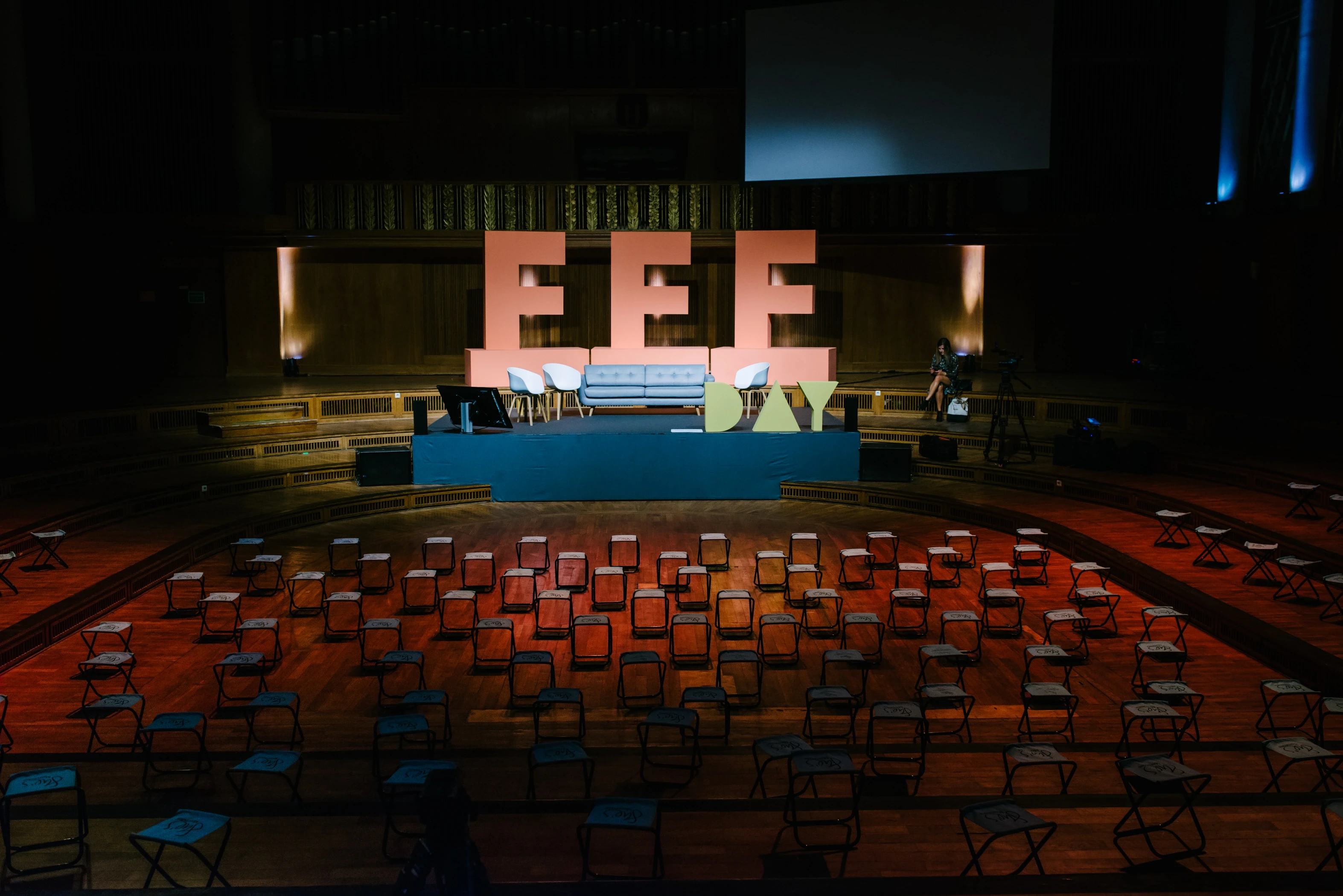 Stage of the FEMALE FUTURE FORCE DAY with oversized coral FFF letters and green DAY lettering, a blue sofa and rows of folding stools in a wood-paneled hall.