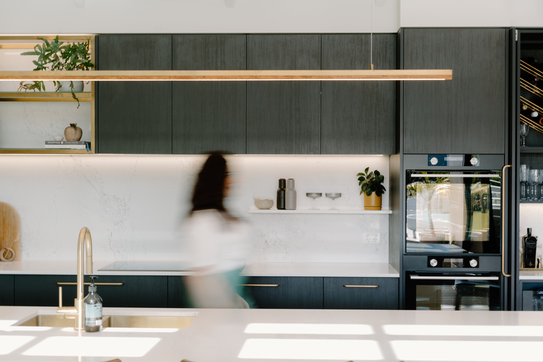 A green, white and gold minimalistic, architecturally designed kitchen with a women walking behind the kitchen bench, blurred