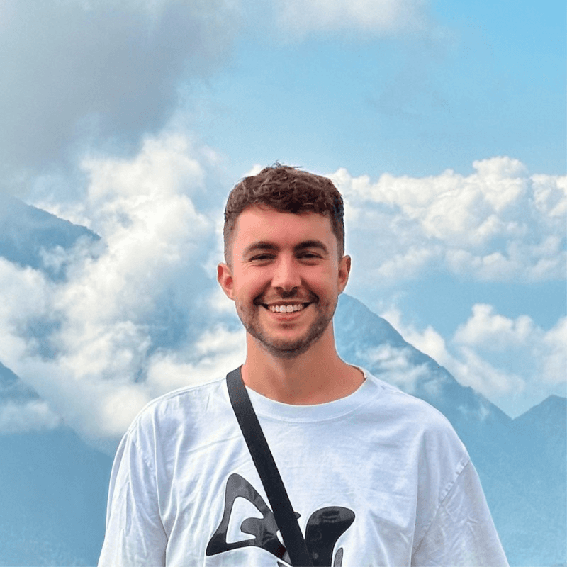 Person wearing a blue puffer jacket and red backpack standing in front of a light blue building on a city street.