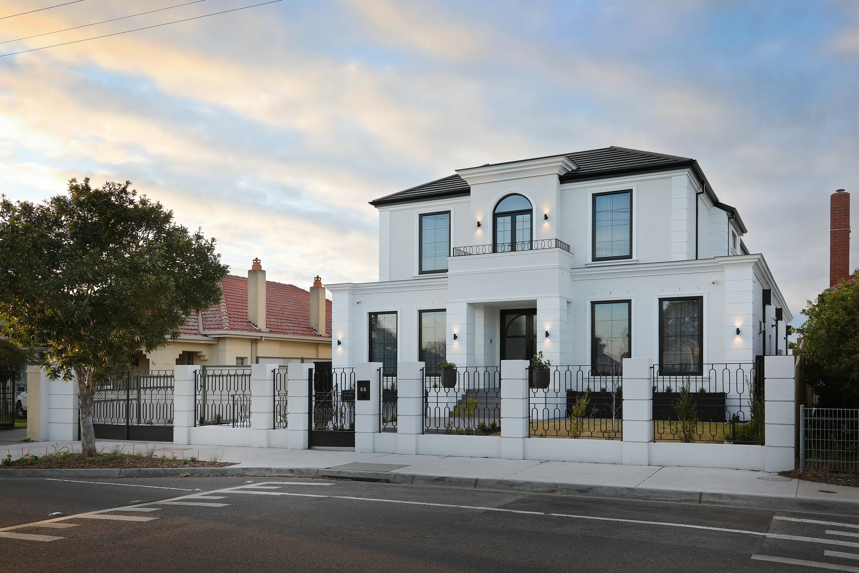 Front facade of the landmark Coburg home at dusk, a rendered two-storey residence with a hipped roof, arched feature window, iron balustrade balcony, columned portico and an ornate iron and white-rendered fence numbered 68