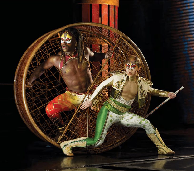 Colorfully dressed characters of KA by Cirque du Soleil stand in a wicker wheel.