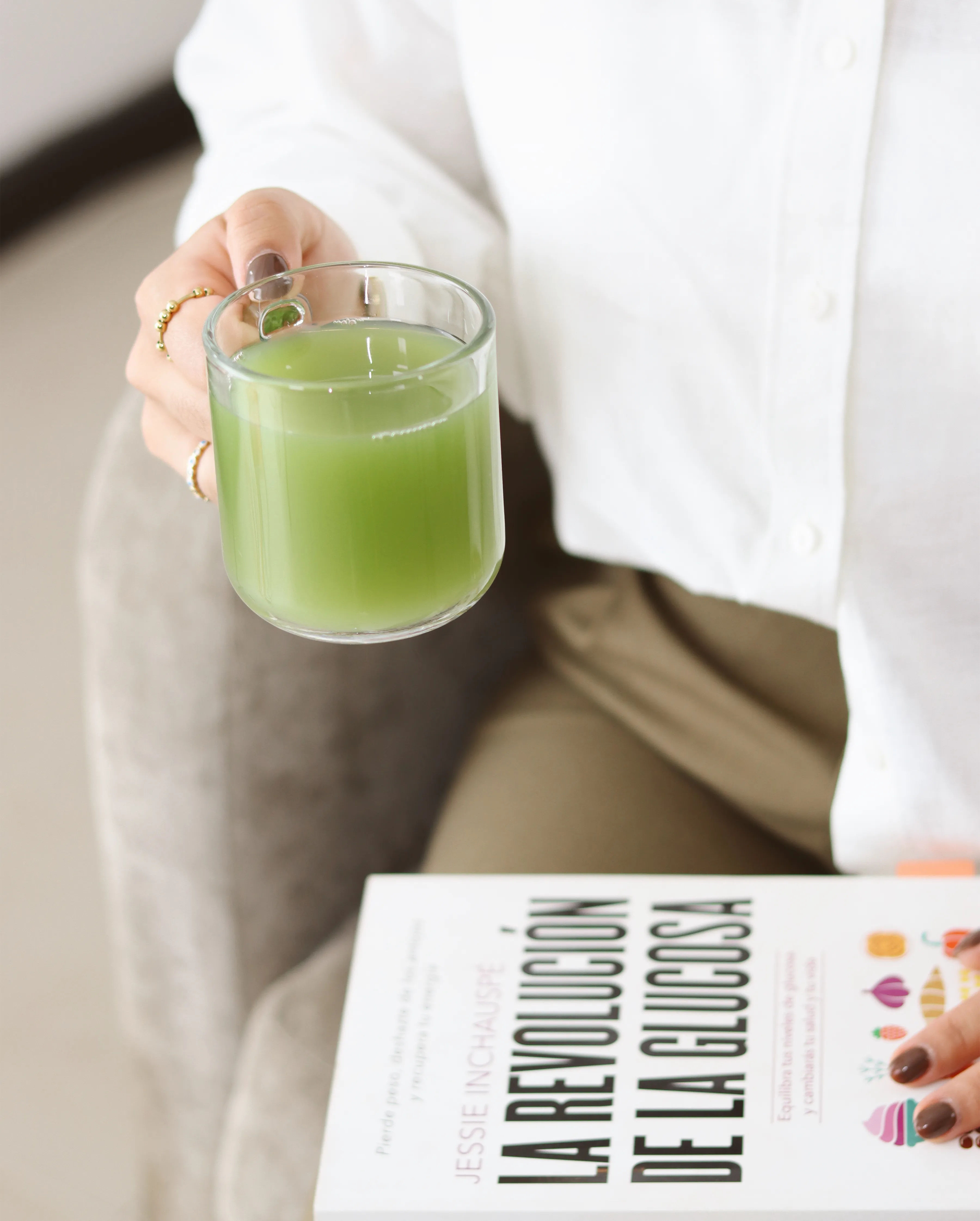 Mano femenina sosteniendo vaso de jugo verde sobre libro.