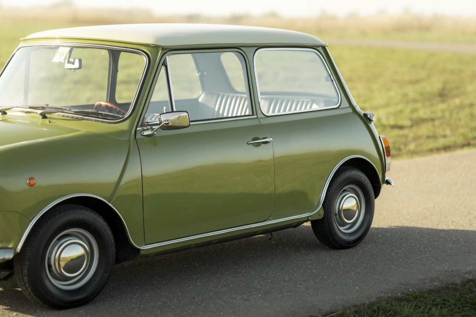 A vintage green car parked on a country road, surrounded by grassy fields under a clear sky.
