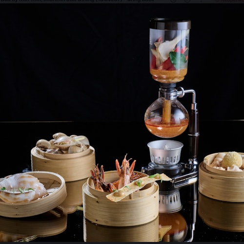 一個虹吸咖啡機，上面擺放著各種菜餚，包括海鮮、餃子和蔬菜，都放在竹蒸籠裡，背景是反射的表面。