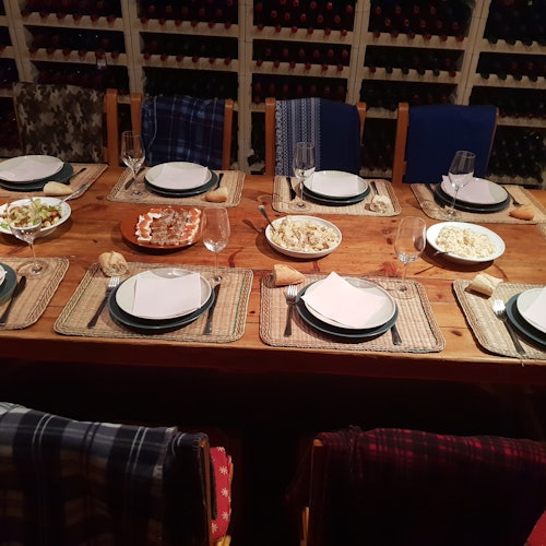 Puinen ruokapöytäsetti kuudelle sisältäen lautaset, aterimet, lasit ja useita kulhoja ruokaa. Taustalla näkyy viinipullotelineitä.