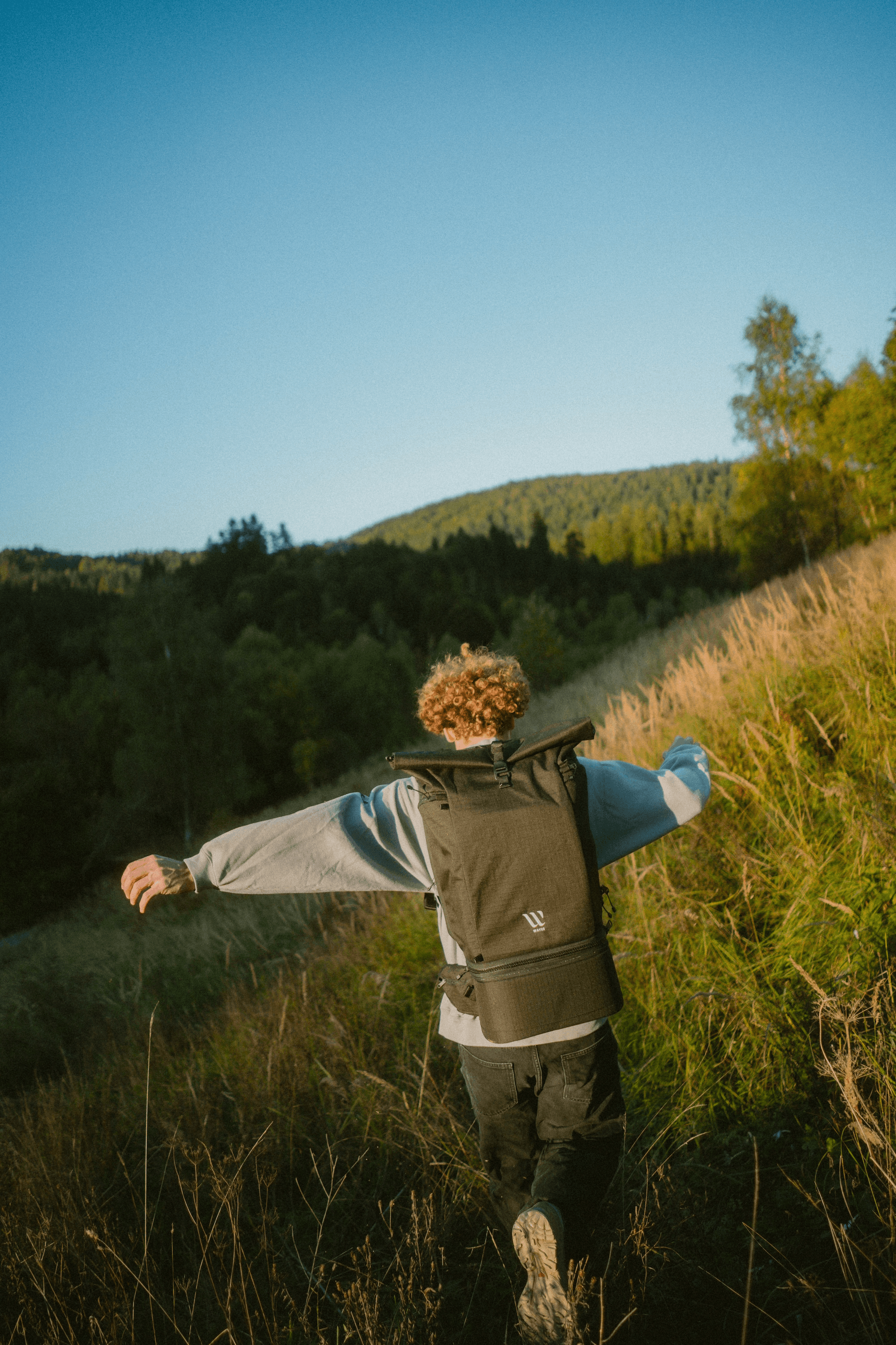 A person with curly hair and a backpack joyfully strides through a grassy hill under a clear blue sky, surrounded by lush, green trees and distant rolling hills.