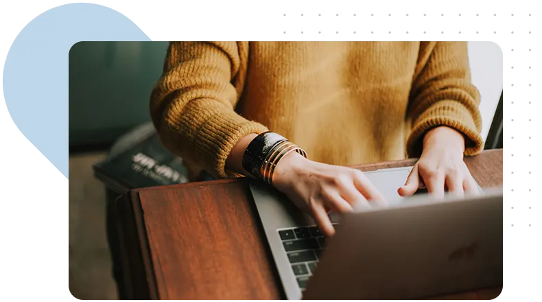 A person in a mustard sweater is working on a laptop, seated at a wooden desk with a cozy atmosphere.