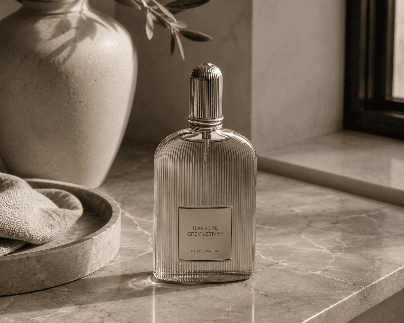 A ribbed translucent glass bottle of Tom Ford Gray Vetiver Eau de Parfum featuring a metallic silver logo label and a ribbed silver cap. The perfume stands on a polished beige marble countertop alongside a gray marble tray, a soft linen cloth, and a large ceramic vase with olive branches under gentle indoor window light.