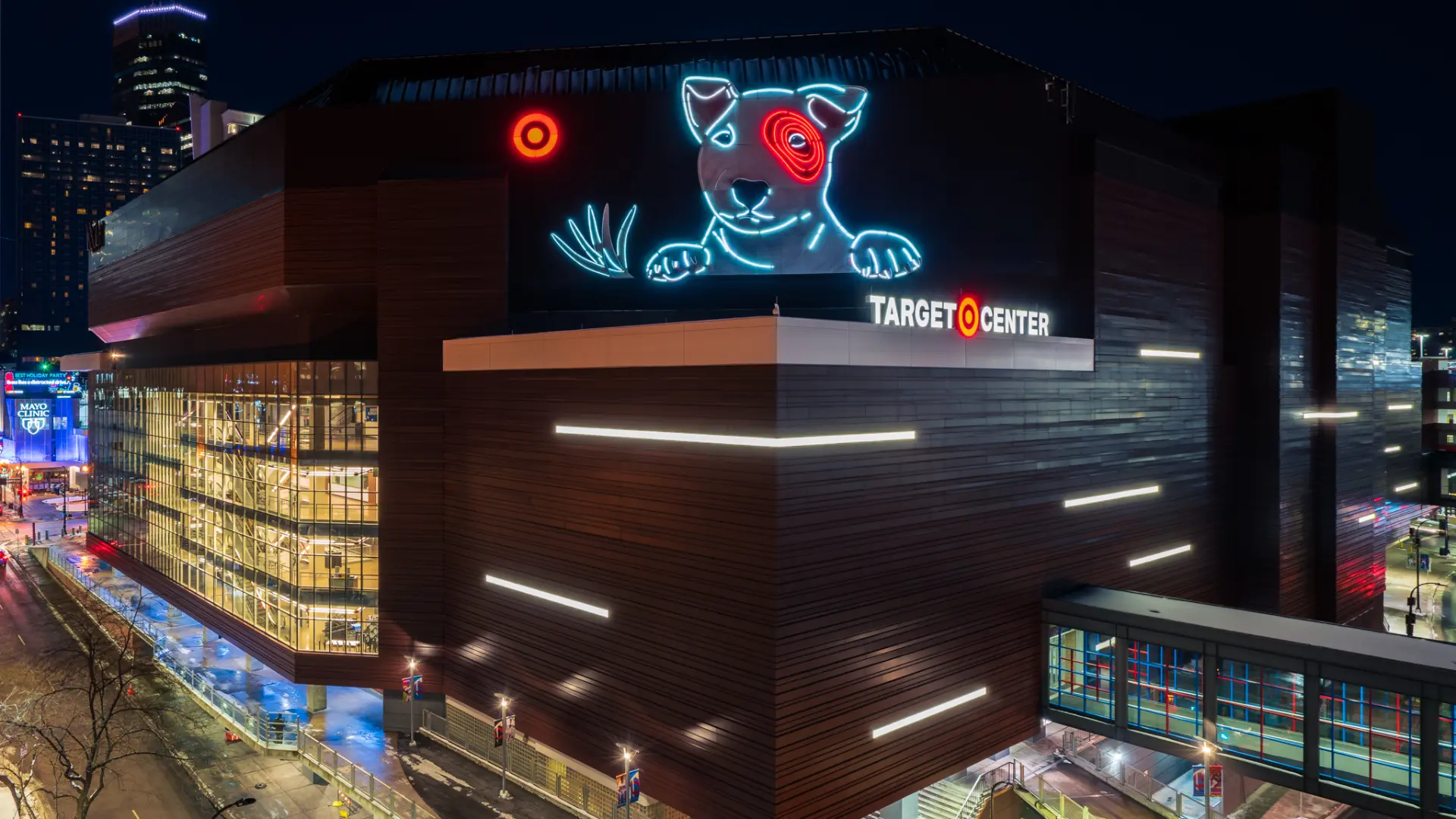 Nighttime Target Center arena glows with a neon bull mural, city lights, wet streets, and busy storefronts, creating an energetic urban mood.