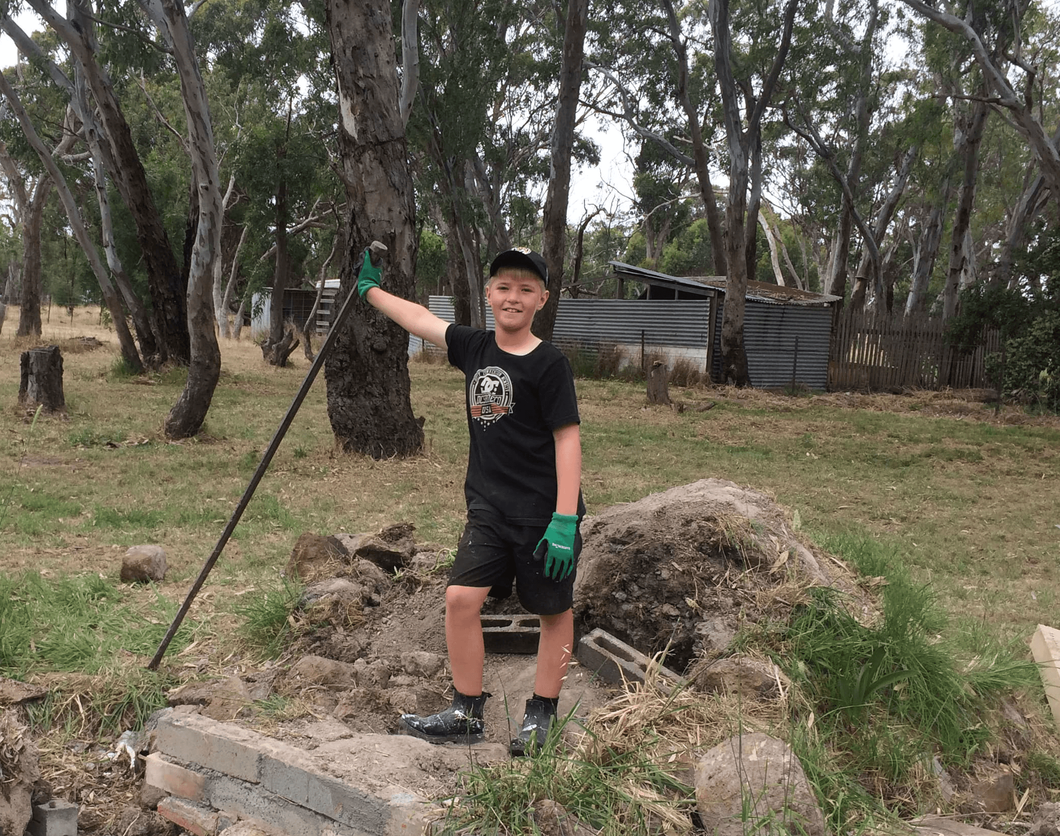 Smiling boy in a black cap and green gloves holds a crowbar.