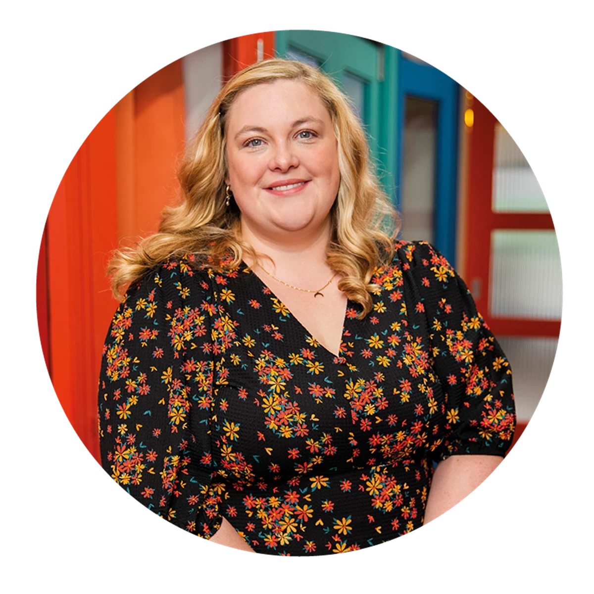 Person wearing a black dress with a colorful floral pattern, standing indoors in front of bright red and blue door frames.