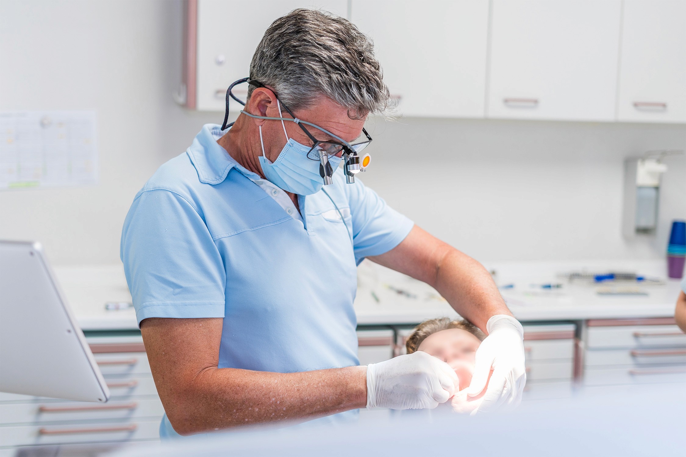 Zahnarzt Dr. Wimmer mit Schutzbrille und Mundschutz behandelt Patient im Behandlungsstuhl in moderner Zahnarztpraxis.