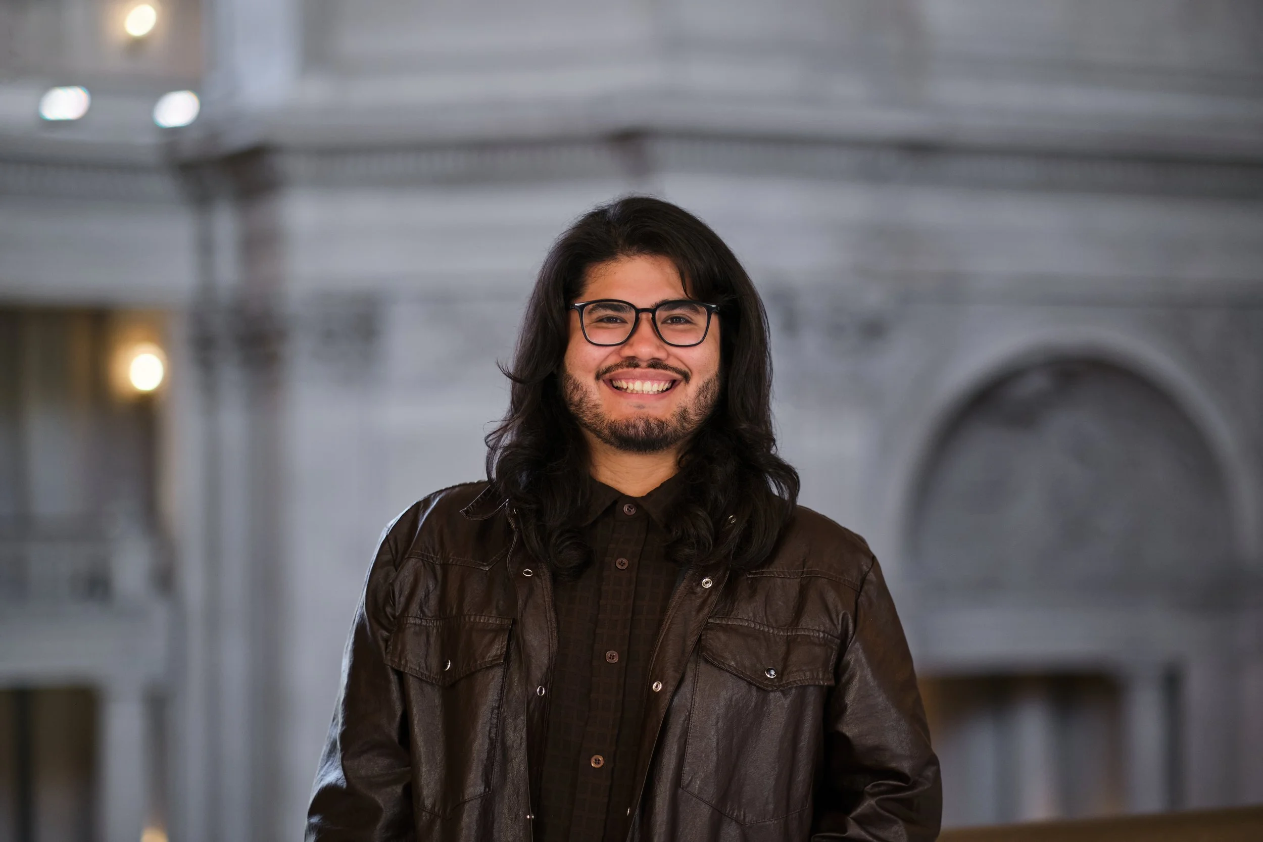 Uma pessoa com cabelos longos e pretos e óculos sorri calorosamente enquanto veste uma jaqueta de couro marrom diante de um fundo arquitetônico ornamentado e desfocado em um ambiente interno, transmitindo uma sensação de confiança e estilo.