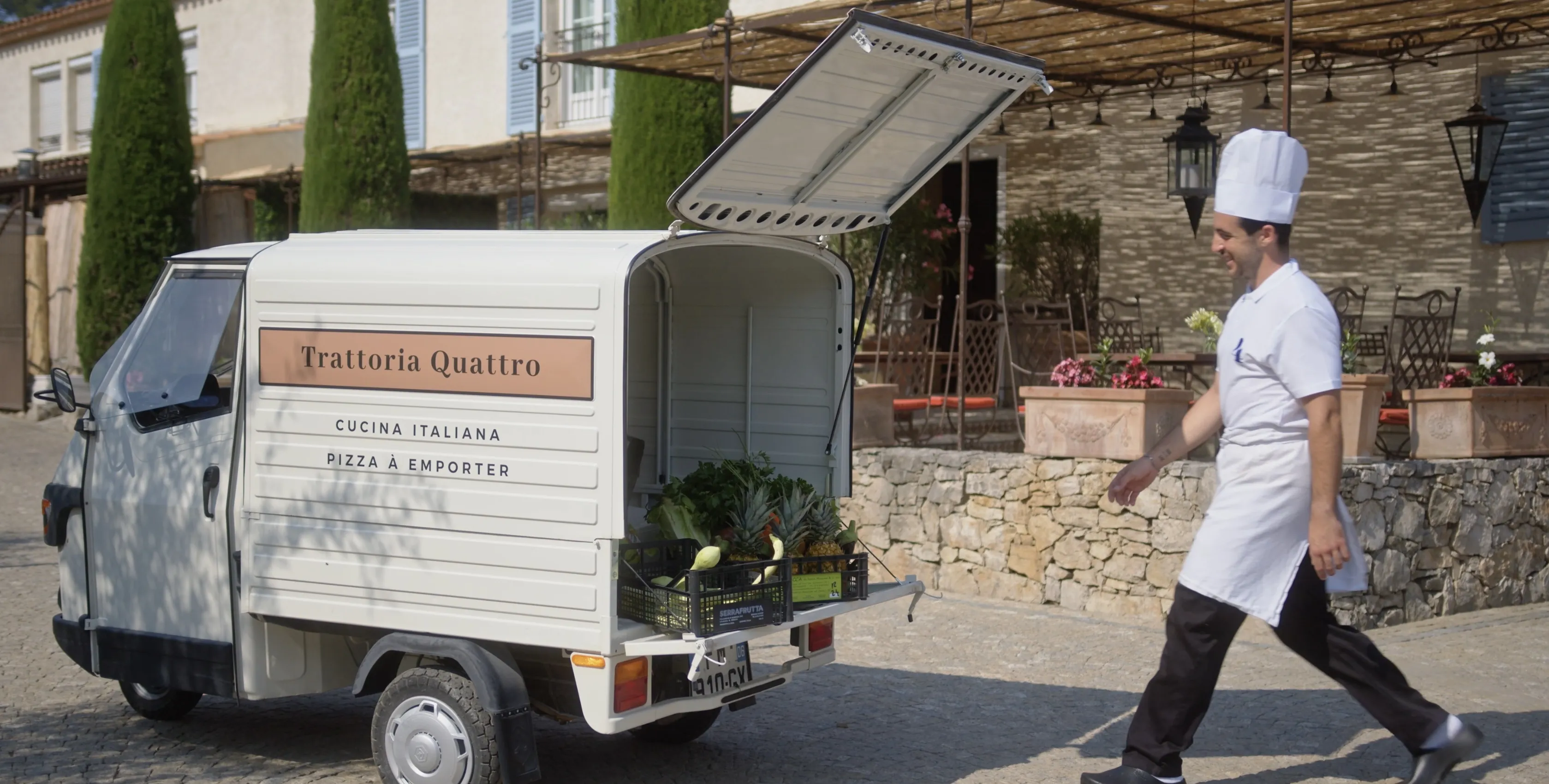 A chef in a white uniform walks by a small delivery truck labeled "Trattoria Quattro," which holds boxes of fresh vegetables, parked on a sunny cobblestone street near a rustic restaurant.