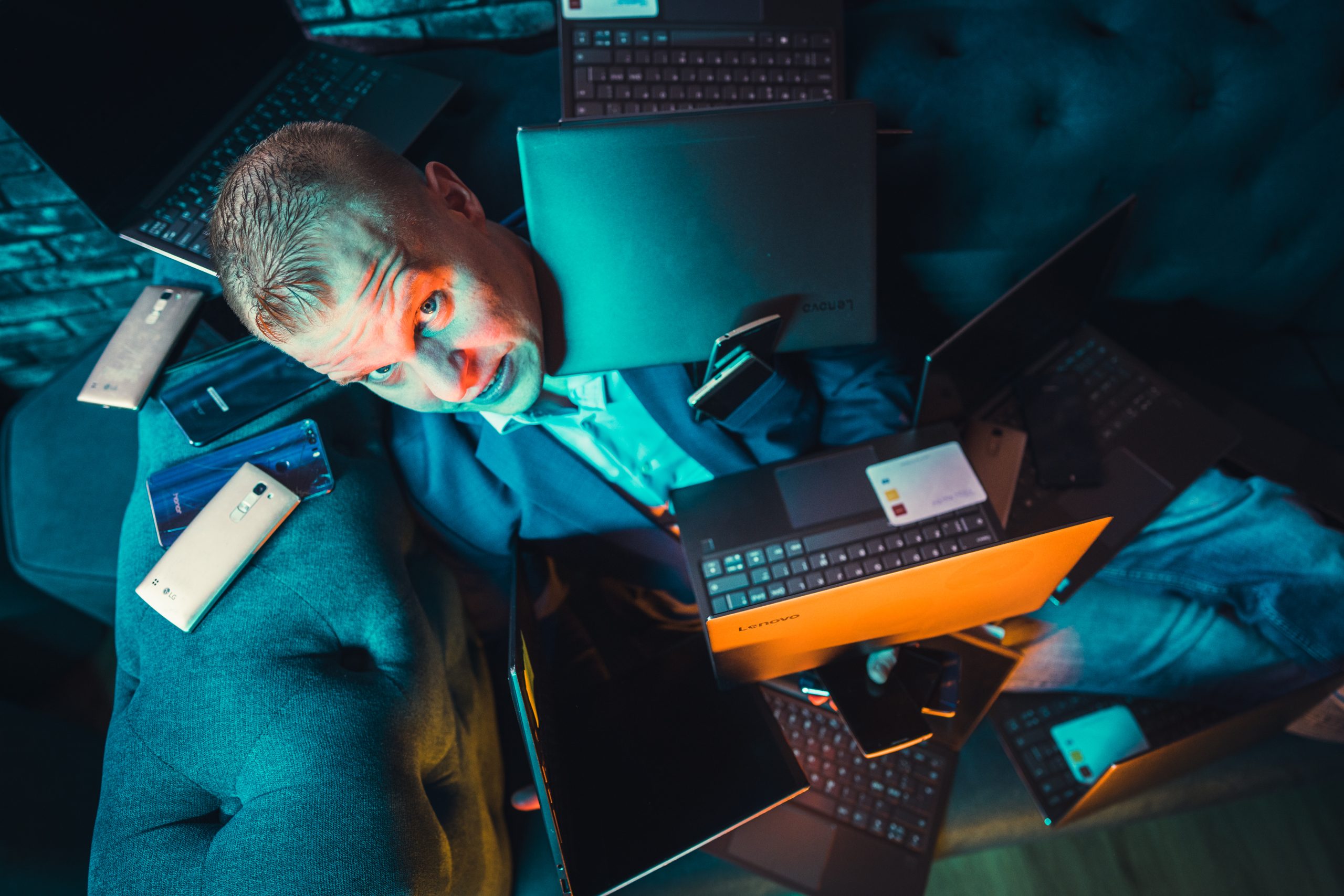 A man in a suit sits surrounded by laptops, smartphones, and credit cards. He looks directly at the camera with a concerned expression