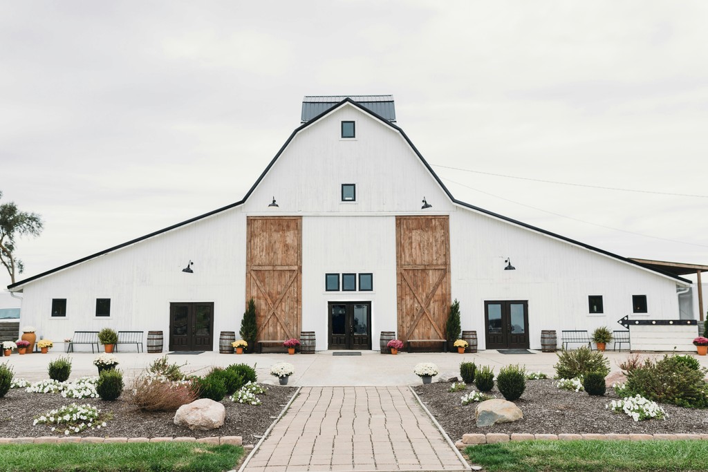 Modern white barn wedding venue with large wooden sliding doors, a paver walkway, and landscaped flower beds with wine barrel accents