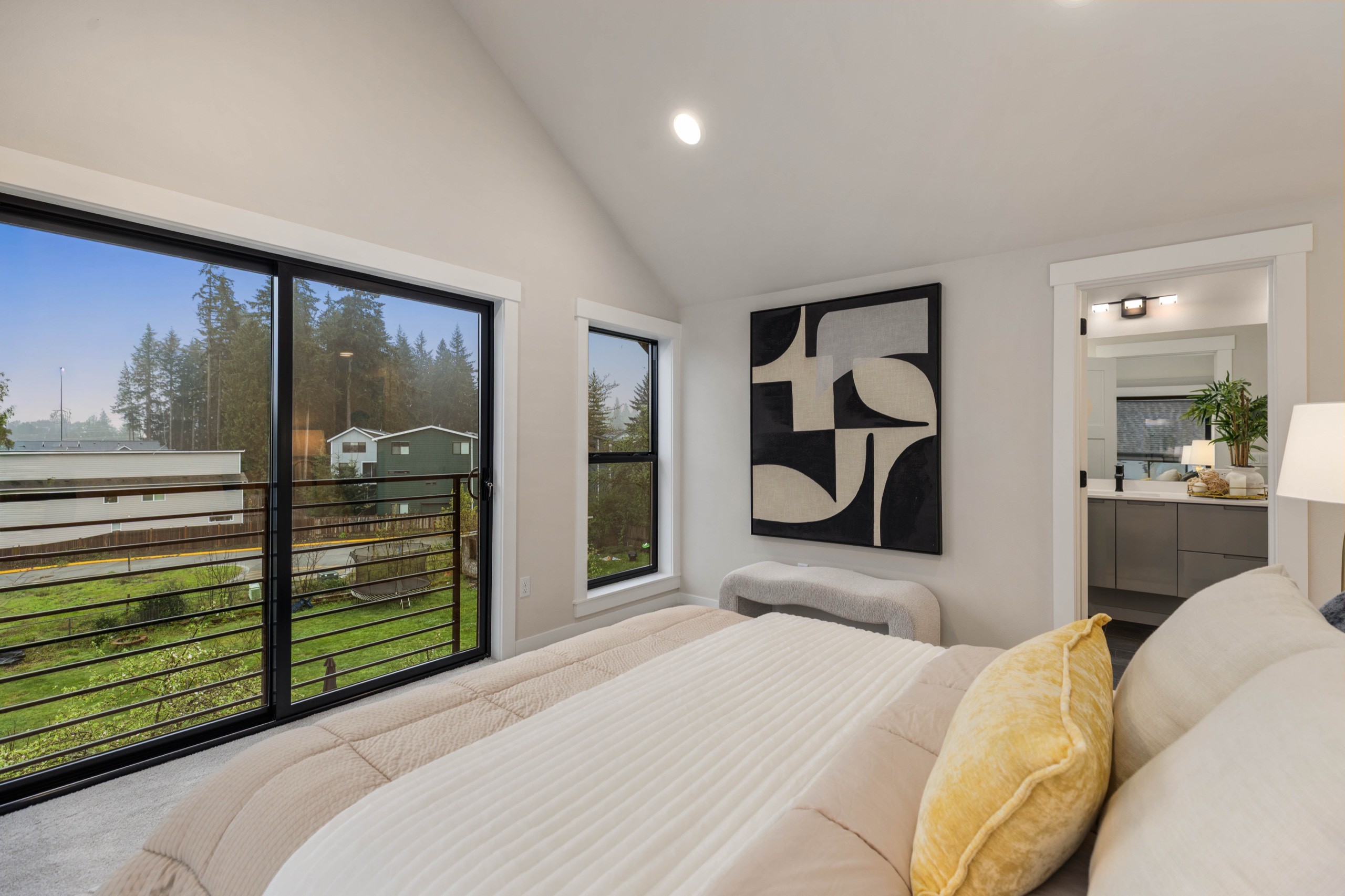 Sunlit luxury bedroom with vaulted ceiling, large sliding glass window overlooking green fields, bold abstract wall art, yellow accent pillows, and ensuite access