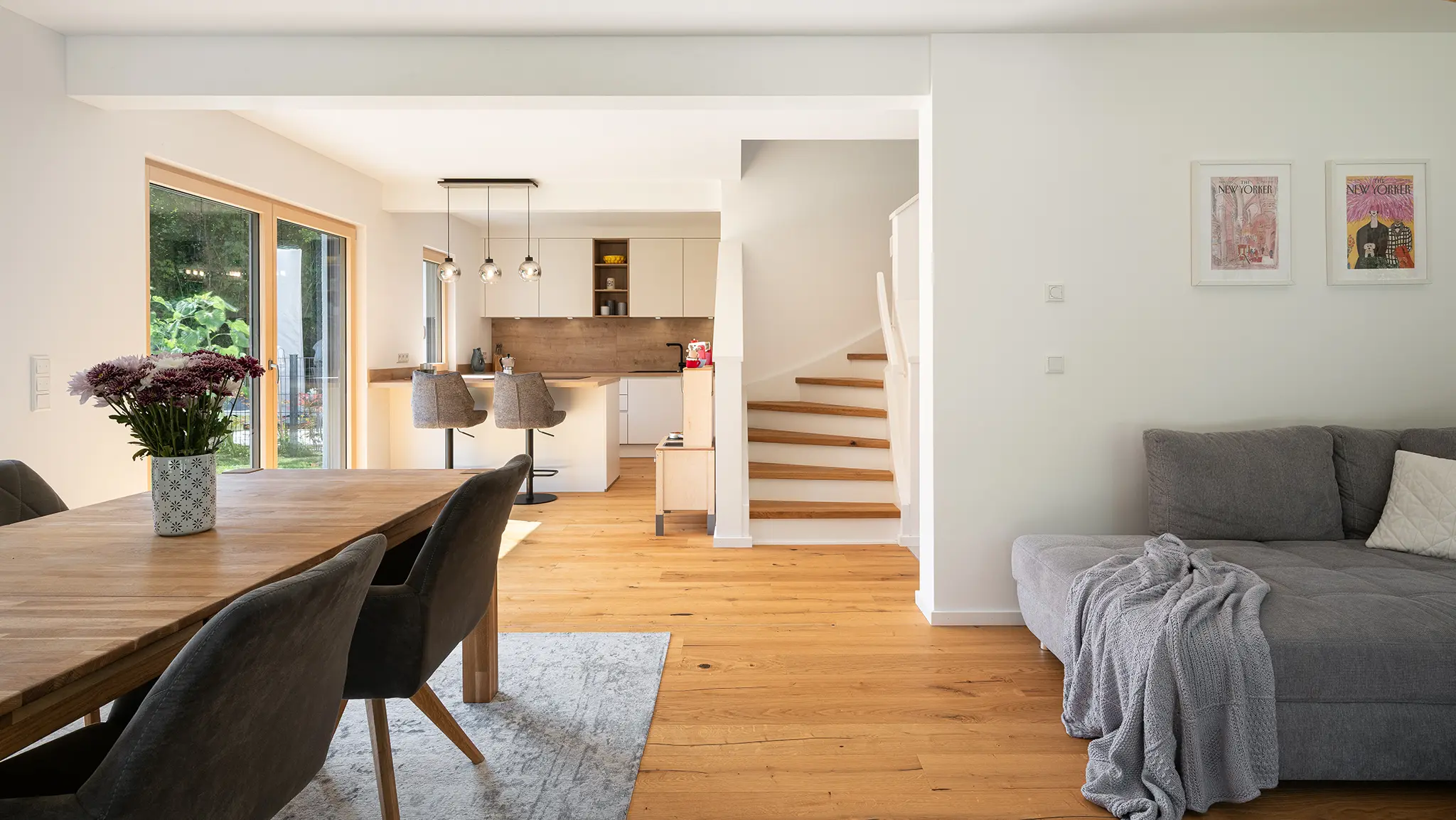 Offener Wohn- und Essbereich mit Holztisch, Stühlen, grauem Sofa mit Decke, Holztreppe und großer Fensterfront mit Blick ins Grüne