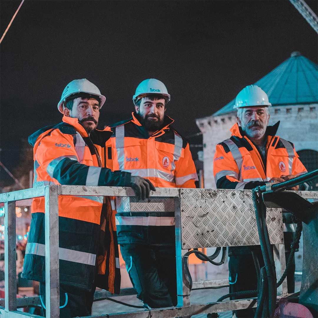 Three male construction workers wearing bright orange coats and blue hard hats, standing together at  a metal railing overlooking a construction site